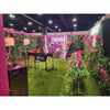 Image of Green Tropics Artificial Plant Wall Panel installed across multiple walls styled with lush greenery, bright pink floral accents, neon signage, spotlights, decorative props, and artificial grass flooring