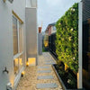 Image of Artificial White Tropics Vertical Garden Wall Panel mounted on a concrete wall in a residential backyard, featuring a stone-paved ground and a glass window