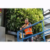 Image of Man in an orange safety vest on a cherry picker with greenery wall in the background