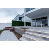 Image of A multi-level modern patio with light-grey stone tiles, LED-lit stairs, a built-in wooden bench, and tall Artificial Boxwood Hedge Plant Wall Screening Panels. White outdoor chairs and glass doors