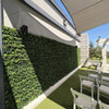 Image of An outdoor event space features the Artificial Boxwood Hedge Plant Wall Screening Panels against a light wall, white folding chairs on artificial turf, a fabric sail overhead, and a white pergola in the background