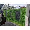 Image of An image of a tall Artificial Deluxe Buxus Hedge Wall Panels that serves as a privacy fence with three black laser-cut panels, bordered by green turf and a gravel driveway in a suburban setting