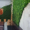 Image of A stylish outdoor patio with Artificial Jasmine Hedge Plant Wall Screen Panels, an exposed red brick accent, rustic wooden seating, and patterned gray and white tile flooring