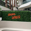 Image of An outdoor setting featuring Artificial Spring Sensation Hedge Wall Panels as a backdrop for a bold orange "APEROL SPRITZ" sign framed by palm trees and curved contemporary architecture