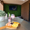 Image of A covered patio with Artificial Spring Sensation Hedge Wall Panels as a backdrop, featuring a mounted TV. In front, a glass table is set with a cheese platter, crackers, grapes, a pink gin & soda, and a large potted plant