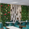 Image of Wild Tropics Artificial Vertical Garden Panels installed indoors, with one decorated for Christmas and the other plain, beside white marble tables and teal chairs separated by a geometric partition wall