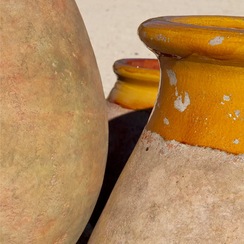 Close-up of rustic ceramic pots with a textured beige body and a glossy mustard-yellow glazed rim, set outdoors on a light sandy surface.