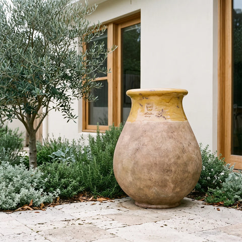 A large rustic ceramic planter pot with a textured beige body and a yellow glazed neck, placed on a stone patio in front of a house with wooden-framed windows and green plants.