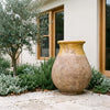 Image of A large rustic ceramic planter pot with a textured beige body and a yellow glazed neck, placed on a stone patio in front of a house with wooden-framed windows and green plants.