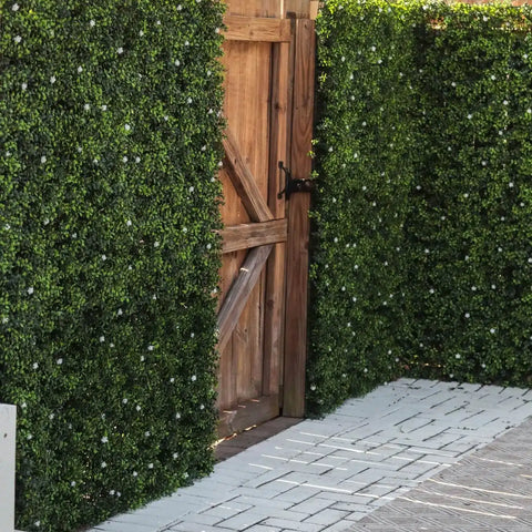 An outdoor scene with a rustic wooden gate framed by tall Artificial Flowering Buxus Hedge Plant Panels with a patterned paver walkway leads to the gate of the garden entrance