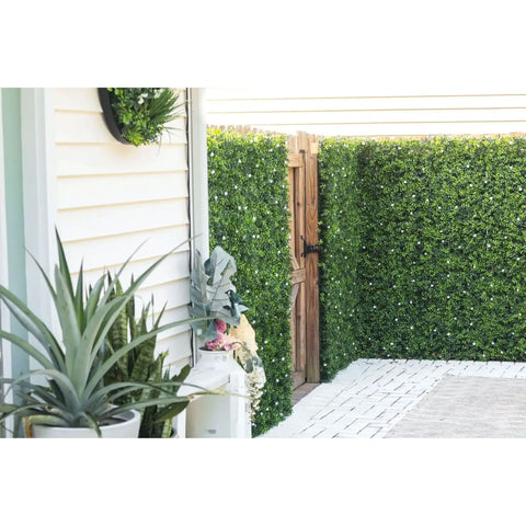 An image of a side yard with the Artificial Flowering Buxus Hedge Plant Panels installed around the rustic wooden gate, white-sided house with wall disc planter, potted greenery, and a clean white paver walkway