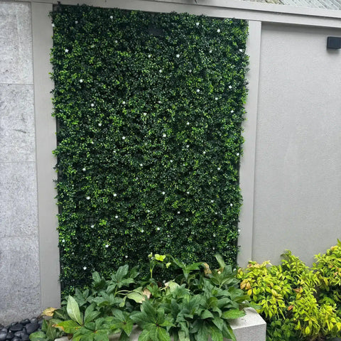 An image of a courtyard featuring the Artificial Flowering Buxus Hedge Plant Panels on a textured gray wall, accented by a planter with lush green and chartreuse plants