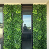 Image of Green Tropics Artificial Plant Wall Panel installed outdoors on both sides of a tall glass window and door 