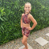 Image of Green Tropics Artificial Plant Wall Panel installed outdoors along a pebbled stone pathway with a gitl in an animal print swimsuit standing in front