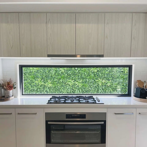 Green Tropics Artificial Plant Wall Panel installed outdoors as a backdrop overlooking the kitchen glass window with stove, oven, potted artificial plants, cooking utensils, and cabinets inside 