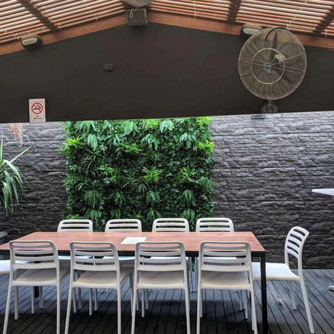 Green Tropics Artificial Plant Wall Panel installed on a brick wall in a patio setting complemented by a shed, an electric wall fan, and dining tables with chairs 