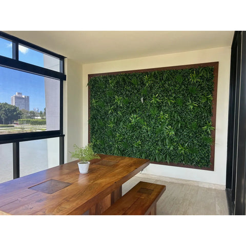 Green Tropics Artificial Plant Wall Panel mounted on a large wood framed wall in  an indoor dining area, paired with a natural wooden table, benches, a small potted plant, and a large window view