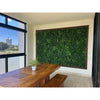 Image of Green Tropics Artificial Plant Wall Panel mounted on a large wood framed wall in  an indoor dining area, paired with a natural wooden table, benches, a small potted plant, and a large window view