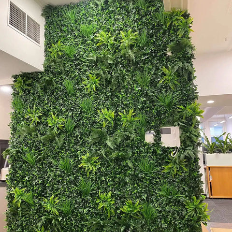Green Tropics Artificial Plant Wall Panel mounted on a large indoor feature wall in a modern office complemented by ceiling lights and nearby workstations