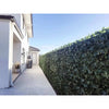 Image of Artificial Peach Leaf Ivy Hedge Screen installed on a backyard fence, set beside a cement pathway with a large potted plant, windows, and a backyard door in view