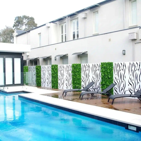 Image of an outdoor pool area with a wooden deck, dark lounge chairs, and a decorative fence alternating black and white patterned panels and Artificial Lavandula Vertical Garden Plant Wall Screening Panels