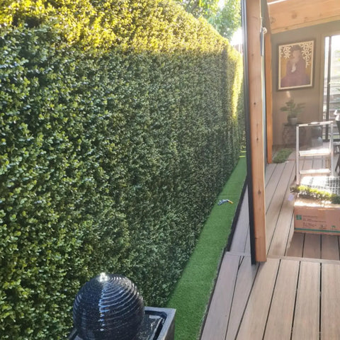 Artificial Premium Natural Buxus Panel installed on a patio with artificial grass beneath and a wooden floor, surrounded by a small ladder, a picture frame, and a pot with an artificial plant