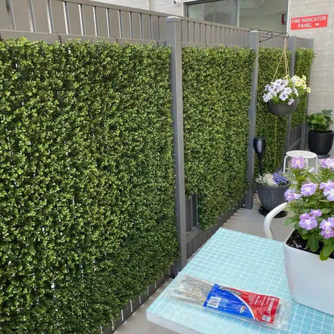 Close up view of the Artificial Premium Natural Buxus Panel installed on a fence, surrounded by various pots containing artificial plants