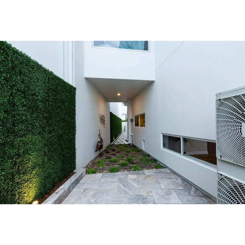 A narrow walkway between modern white buildings, highlighted Artificial Boxwood Hedge Plant Wall Screening Panels, a paved stone path, a small garden bed, and a decorative bird sculpture