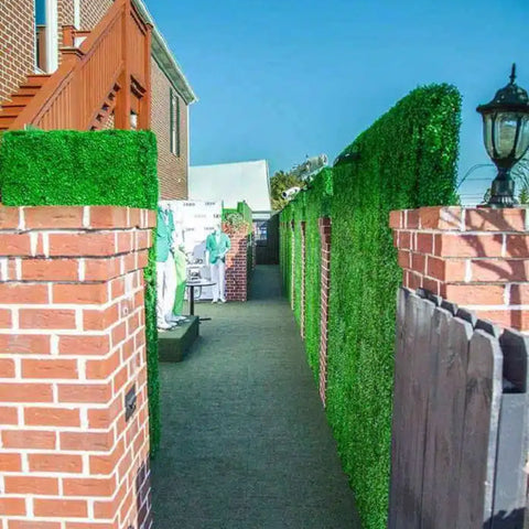 A narrow outdoor walkway with Artificial Boxwood Hedge Plant Wall Screening Panels and brick pillars on one side, artificial turf underfoot, and a brick building with a staircase on the other, leading toward a distant reception area