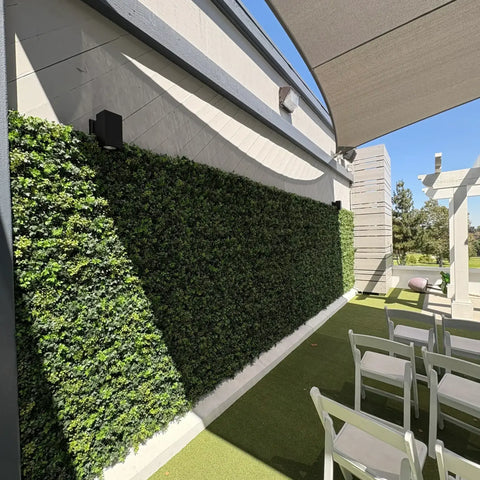 An outdoor event space features the Artificial Boxwood Hedge Plant Wall Screening Panels against a light wall, white folding chairs on artificial turf, a fabric sail overhead, and a white pergola in the background