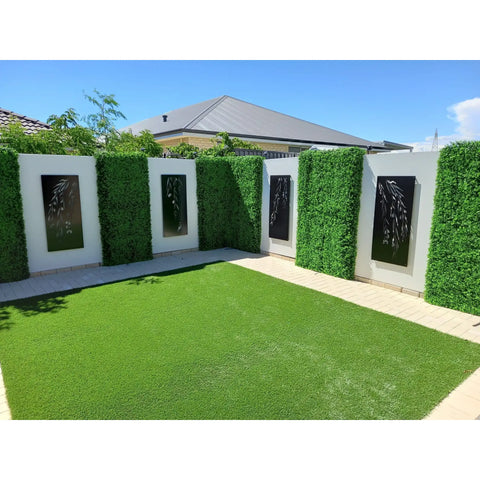 An image of a backyard with patterned walls of alternating Artificial Deluxe Buxus Hedge Wall Panels and laser cut black panels, bright green turf, light pavers and a home roof under a clear sky