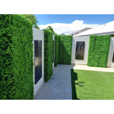 A photo of a backyard walkway with light pavers, patterned walls of Artificial Deluxe Buxus Hedge Wall Panels and laser-cut black panels and bright green turf under strong sunlight