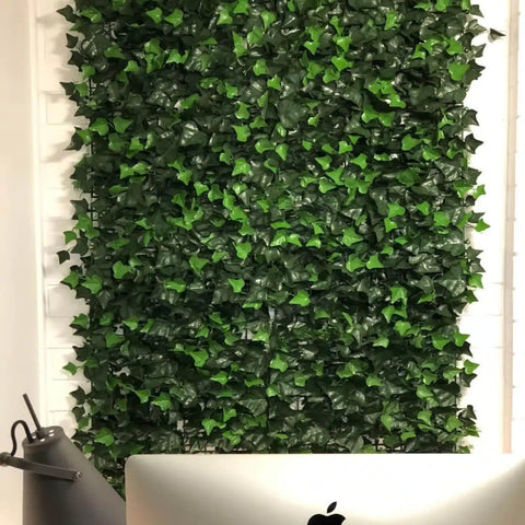 An image of a workspace featuring the Artificial Ivy Leaf Hedge Panel as a green backdrop behind a desk with a silver Apple monitor and minimalist dark gray lamp