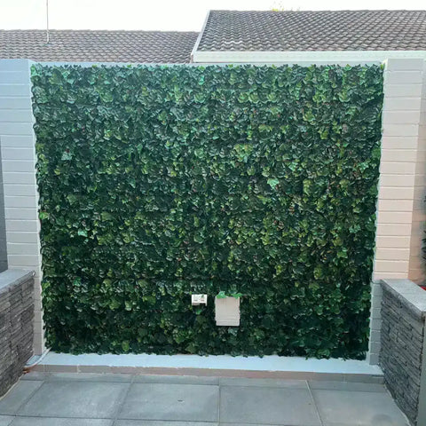 An image of a large Artificial Ivy Leaf Hedge Panel installed on an outdoor feature wall, framed by light gray brick pillars and contrasting with gray square tiles below with precise cuts around a small utility box