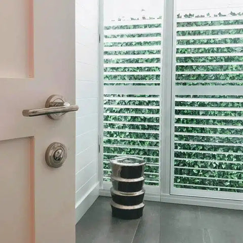 A room corner featuring white plantation shutters. The partially open slats look out onto the Artificial Ivy Leaf Hedge 1m x 1m Plant Wall Screening Panel UV Protected installed on a wall outdoors
