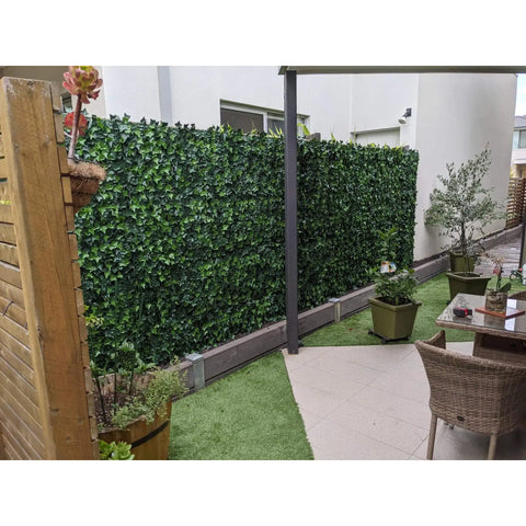 Image of a modern backyard patio featuring a tall Artificial Ivy Leaf Hedge Panel that acts as a lush green privacy screen. The space is divided between a paved area with dining set and an adjacent patch of bright artificial grass