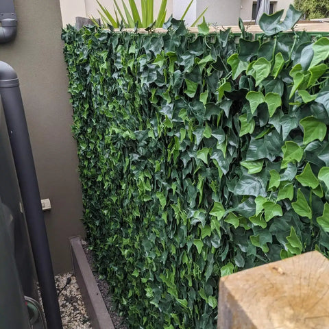 Close up view of the Artificial Ivy Leaf Hedge Panel installed in a narrow outdoor passage. The ground is covered with gravel with a large utility pipe on the left and a thick wooden fence post is visible in the immediate foreground