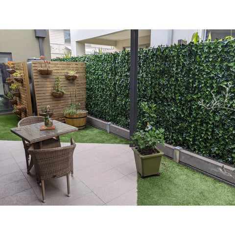 A private courtyard featuring the Artificial Ivy Leaf Hedge Panels, wooden slat screen with hanging planters, tiled patio with dining set, potted plants, and a mix of tiles and artificial turf