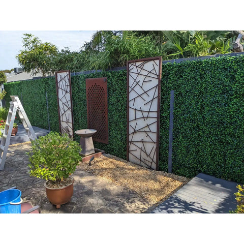 A landscaped backyard with a decorative Artificial Jasmine Hedge Plant Wall Screen Panels featuring three art panels, a gravel bed with a stone water feature, flagstone patio, dark deck, a ladder, and potted plants
