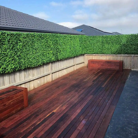 A modern backyard with a reddish brown wooden deck, tall Artificial Jasmine Hedge Plant Wall Screen Panels, matching storage benches, and neighboring rooftops in the background