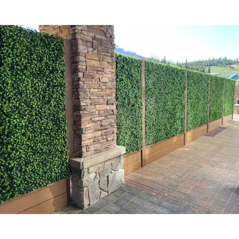 Image of Artificial Jasmine Hedge Plant Wall Screen Panels mounted with wood look framing and a stacked stone pillar on a paved patio, set against a tree lined mountain slope