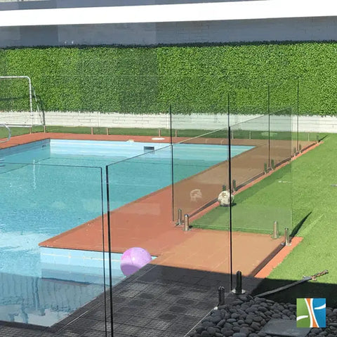 An image of a backyard pool with reddish brown deck and coping, artificial turf, Artificial Jasmine Hedge Plant Wall Screen Panels that serves as a privacy wall, glass safety fence, and a floating purple ball, highlighted by the sunlight
