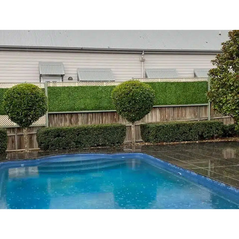 A landscaped pool area on a rainy day, featuring dark tiled decking and a layered privacy fence with topiary trees, wooden panels, lattice, and Artificial Jasmine Hedge Plant Wall Screen Panels