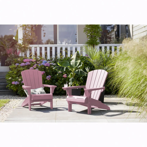 Two pink Adirondack chairs on a patio with plants and flowers in the background.