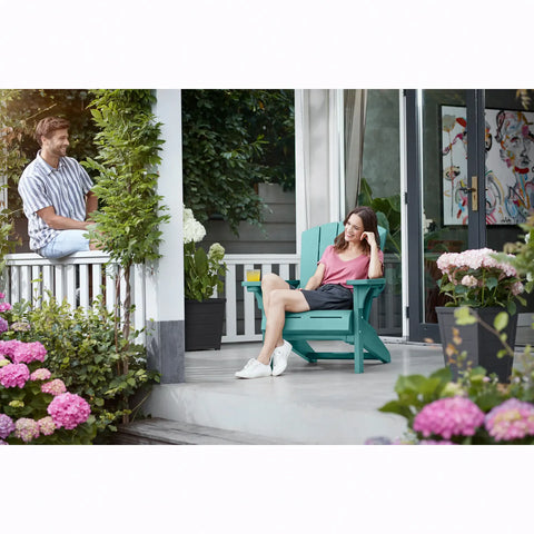 Two people sitting on a porch with teal Adirondack chairs, surrounded by flowers and plants.