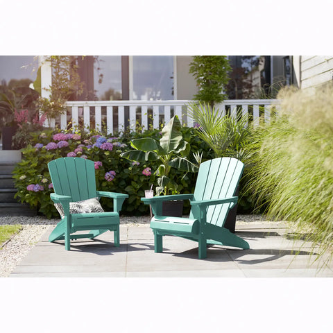 Two teal Adirondack chairs on a patio with plants and flowers in the background.