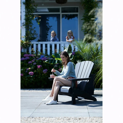 Woman sitting on a graphite Adirondack chair in a garden setting with flowers and a house in the background.