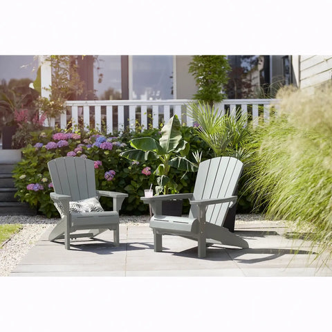 Two gray Adirondack chairs on a patio with plants and flowers in the background.