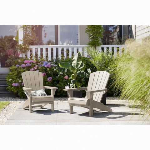 Two Adirondack chairs on a patio with plants and flowers in the background.