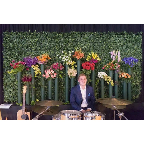 A young man smiles behind a drum set in front of dense Artificial Laurel Hedge Plant Wall Screening Panels adorned with colorful flower bouquets, with an acoustic guitar and a drum set nearby